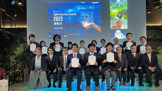 Cybersecurity Awards 2023 表彰式のステージで撮影された集合写真。背景のスクリーンにはイベント名と日付（2023年3月3日開催）が表示され、右側にはオンライン参加者の映像も映し出されている。ステージ上には受賞者や関係者が複数列に並び、中央列の人々は賞状を持っている。